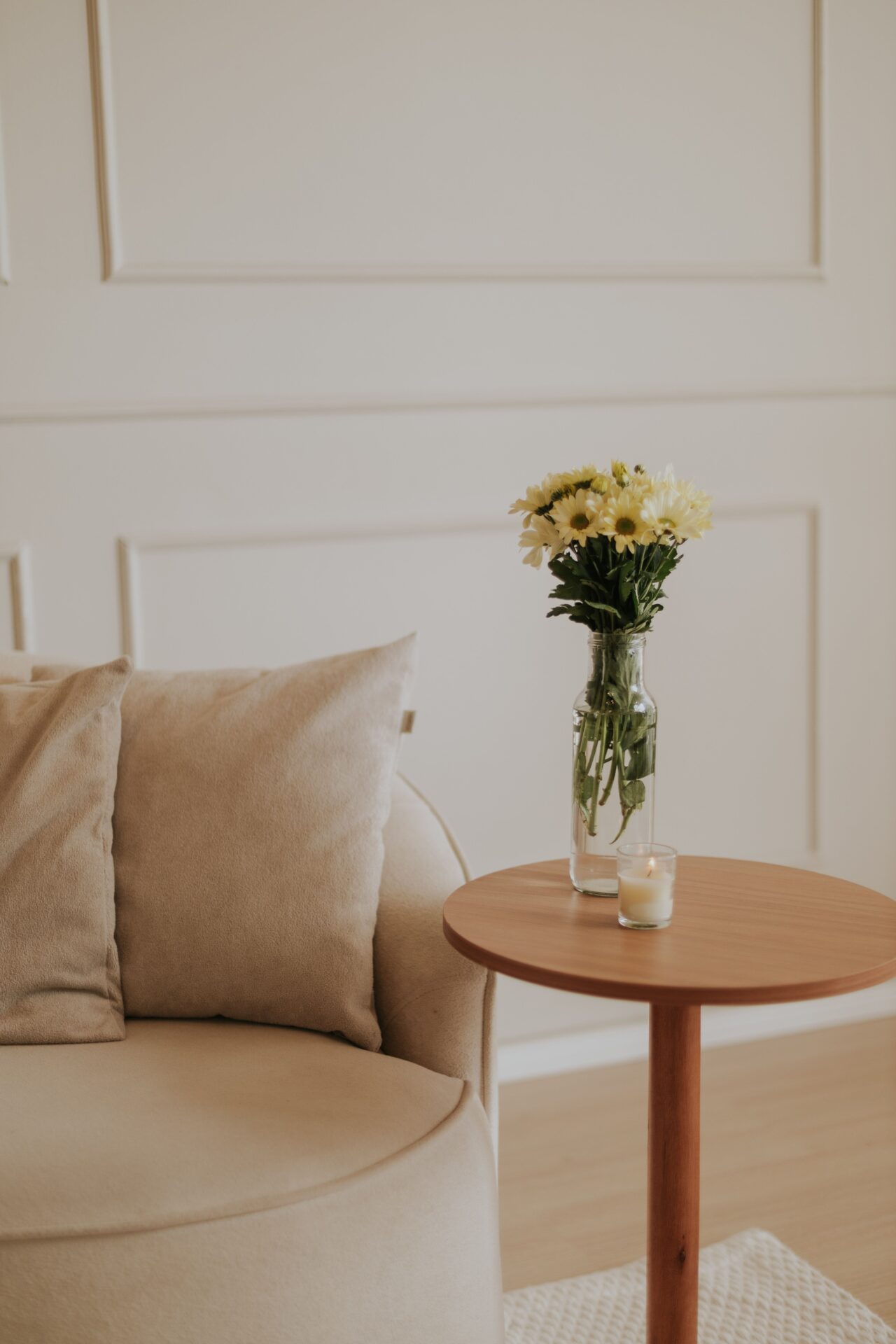 Neutral upholstered chair beside a round wood table holding yellow flowers.
